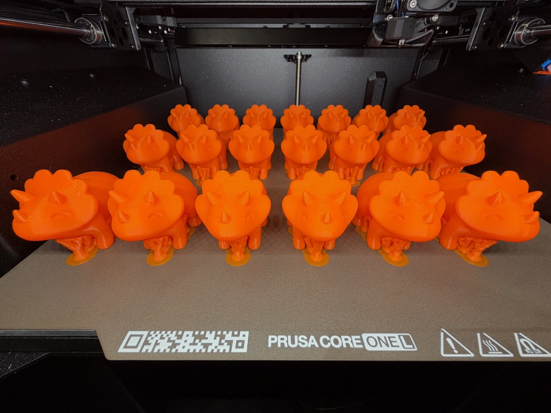 20 orangene Triceratops-Modelle auf einem Druckblech in einem Prusa CORE One L